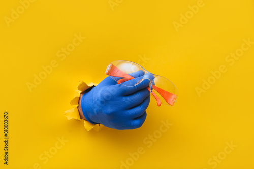 The right male hand in a blue fabric work glove holds orange safety glasses. Torn hole in yellow paper. Good job, eye protection and safety concept. Copy space. photo