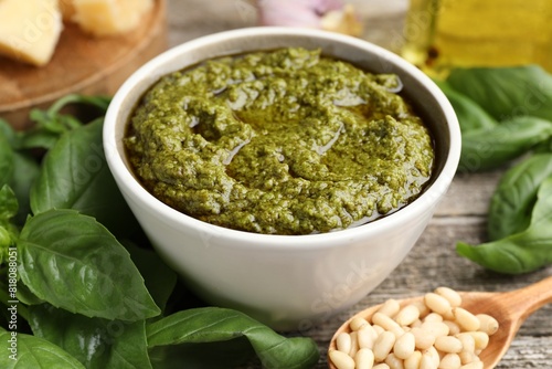 Tasty pesto sauce in bowl, pine nuts and basil on wooden table, closeup