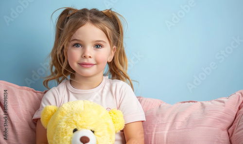 Smiling Young Girl Holding Yellow Teddy Bear Sitting on Pink Couch Against Blue Background