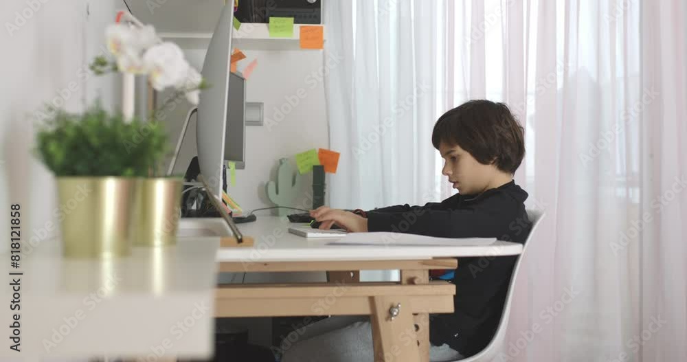 A young boy focused on his homework while working on a laptop during daylight hours. Post-it notes and school supplies are visible on the desk. A young boy focused on his homework while working on a laptop during daylight hours. Post-it notes and school supplies are visible on the desk.