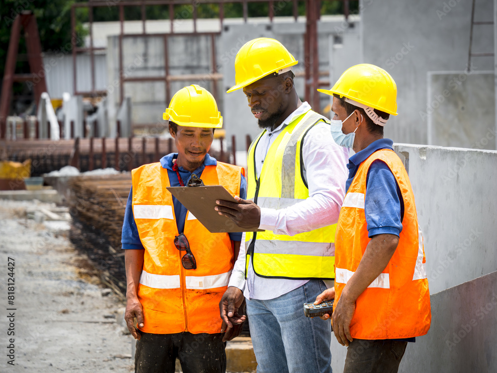 African male foreman and two Asian workers in safety vests and helmets stand by a building under construction, one holding a clipboard, pointing and explaining. African male foreman and two Asian workers in safety vests and helmets stand by a building under construction, one holding a clipboard, pointing and explaining.