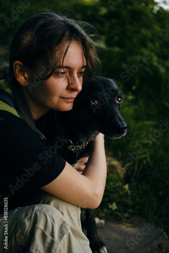 Young woman hugs with black mix breed dog in the middle of a green spring park. A dog with female owner rests outside on a summer sunny day in shadows of trees.