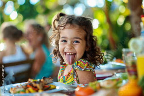 Cheerful little girl sitting at a table outdoors, indulging in grilled food at a family garden gathering, playfully sticking her tongue out.