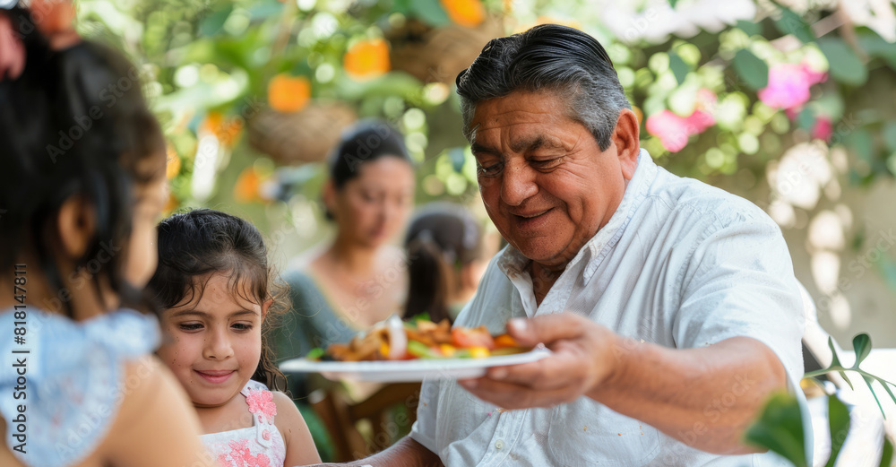 Latino man offering a plate of food to a little girl seated at a table. Dining on grilled food outdoors during a family garden party.