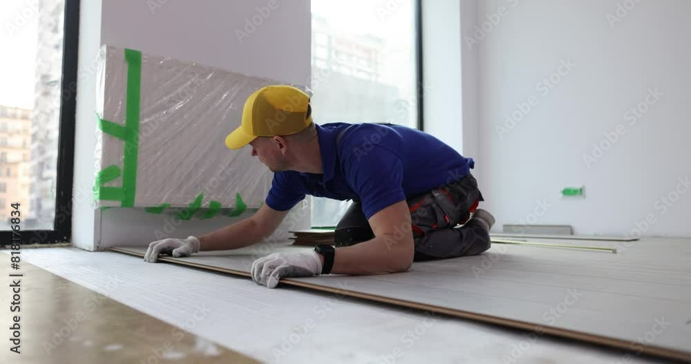 Worker with cap installs laminate board on floor in office enclosure. Employee lays laminate in repaired apartment. Skilled builder at work