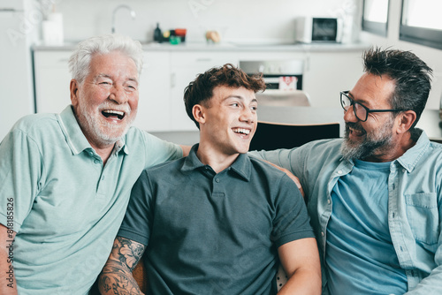Happy multigenerational family of males stay together with fun. Grandfather, mature son and young grandson laughing while sitting on the sofa at home