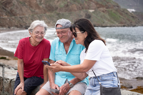Group of senior Caucasian friends in leisure travel at sea sitting looking together at mobile phone. Three seniors smiling people enjoying retirement and free time in vacation #818186250