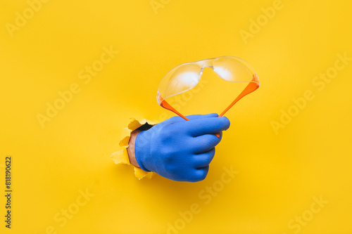 The right male hand in a blue fabric work glove holds safety glasses. Torn hole in yellow paper. Good job, eye protection and safety concept. Copy space. photo