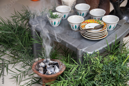 A jebena sits on a grass-covered table with small, handle-less cups arranged around it. Roasted coffee beans, a mortar and pestle, and incense smoke create a traditional Ethiopian coffee ceremony
