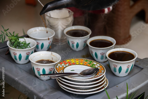 A jebena sits on a grass-covered table with small, handle-less cups arranged around it. Roasted coffee beans, a mortar and pestle, and incense smoke create a traditional Ethiopian coffee ceremony