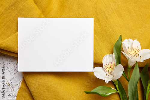 Top view of a landscape-oriented white card on mustard-colored fabric, complemented by white Alstroemeria flowers © mikeosphoto