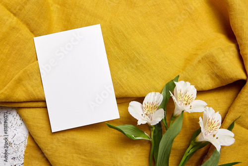 An upright white card on a rich mustard-yellow cloth, accented by white Alstroemeria flowers, flat lay © mikeosphoto