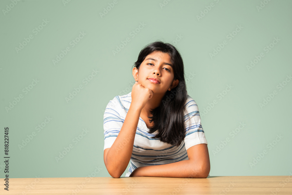 Indian asian small girl sitting at table against plain green background Indian asian small girl sitting at table against plain green background