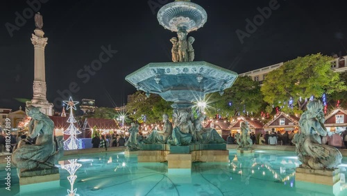 Panorama showing illuminated fountain with evening timelapse view of holiday decorations and food booths at the Rossio Christmas Market on Dom Pedro IV square in Baixa district of Lisbon, Portugal.