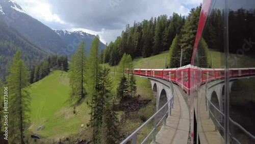 Grisons, Switzerland - April 12. 2024: Bernina Express, red train pass over a old stones bridge with arches in beautiful Swiss Alps landscape photo