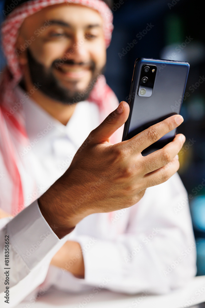 Middle Eastern entrepreneur in traditional clothing sits at a modern desk, browsing the internet on his cell phone. With a smile, an Islamic man uses wireless technology for work. Close-up shot. Middle Eastern entrepreneur in traditional clothing sits at a modern desk, browsing the internet on his cell phone. With a smile, an Islamic man uses wireless technology for work. Close-up shot.