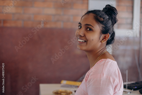 Portrait of charming young Indian woman in pink dress smiling at camera inside room. Positive. 