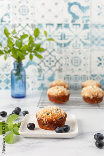 One Homemade Blueberry Muffin on a White Plate with More Muffins  on a Cooling Rack in the Background in a Blue and White Kitchen