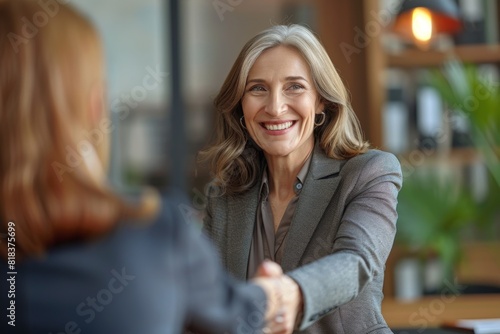 A smiling businesswoman greets another professional with a handshake in a cozy office environment