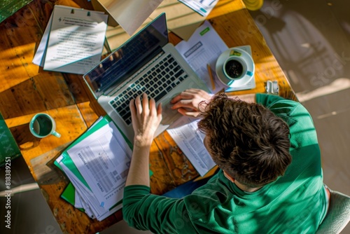Man in a green shirt working on her laptop with a coffee cup and papers, from a high angle view, in the style of professional photography. photo