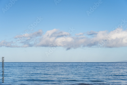 Peaceful, serene, and calming blue nature background of flat Pacific Ocean reaching to a blue sky horizon with line of white fluffy clouds, as a nature background 