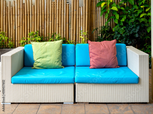 Empty white modern rattan couch seat with blue cushion, sofa with pillows for guests on bamboo wall background at the outdoor garden with nobody. Asian style summer furniture, relaxation concept.