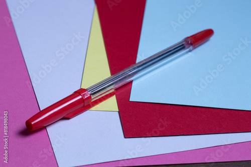Generic red ballpoint pen sitting on several sheets of colored paper.