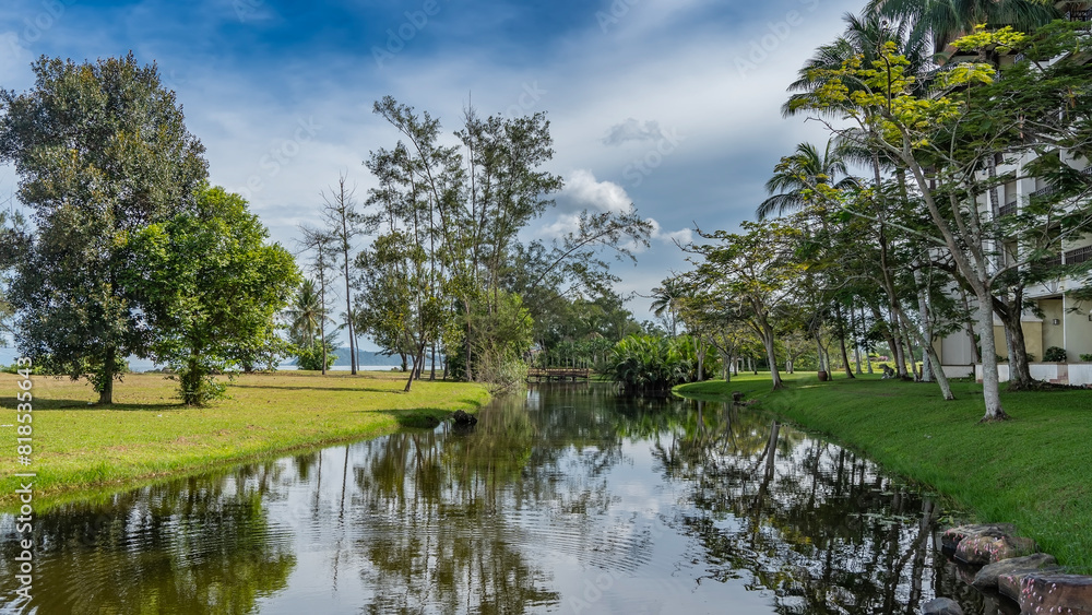 Fototapeta premium A calm river in a tropical park. Sprawling trees grow on the banks. Green grass on the lawns. A fragment of the hotel building in the distance. Clouds in the blue sky. Reflection. Malaysia. Borneo
