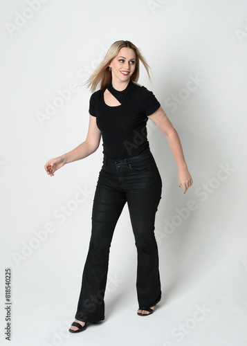 Full length portrait of beautiful blonde woman wearing modern black shirt and leather pants. Confident standing pose with gestural hands presenting, silhouetted on white studio background.