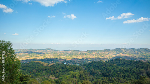 panoramic view of a hilly landscape under a clear blue sky. The foreground features lush greenery, indicating a forested area. As the view extends, rolling hills covered in patches of green and brown 