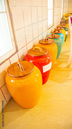 row of colorful ceramic jars against a tiled wall, each jar vibrant and glossy, ranging from warm yellow to cool blue. The jars have wooden lids, adding an earthy touch to the vivid ceramics. A small 
