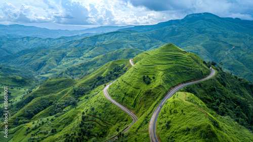 High-altitude view of a curvy mountain road, snaking through lush green hills under a partly cloudy sky,