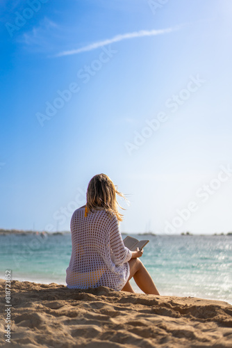 Mid-adult beautiful woman sitting on sunny beach by the sea reading book 