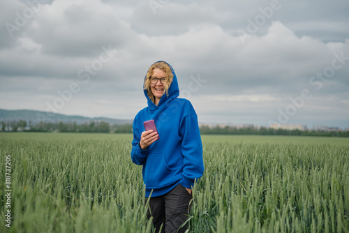 Portrait of a smiling adult blonde woman in a blue sweatshirt with a phone in her hand  portrait against the background of field of young green wheat under gloomy pre-storm cloudy sky  selective focus