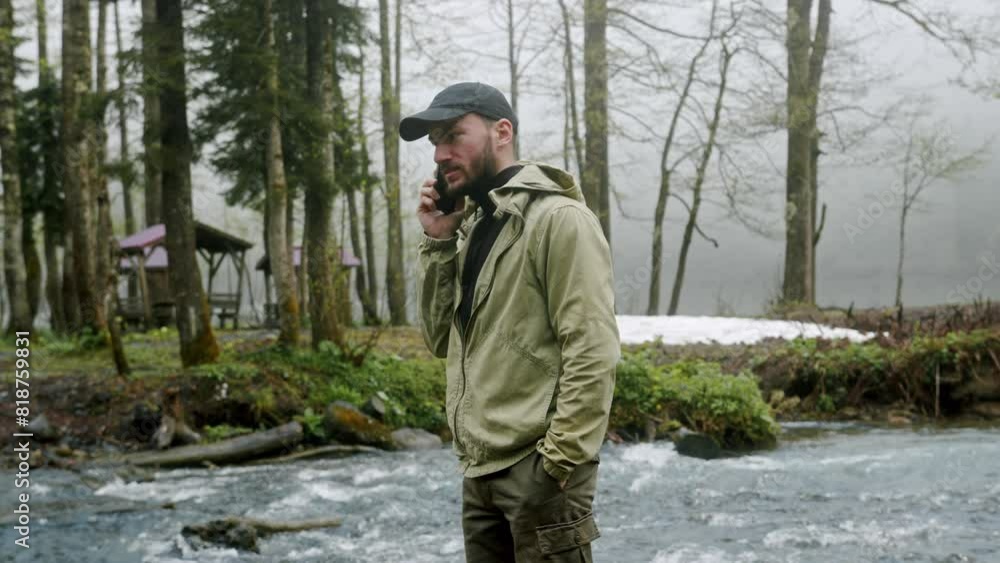 Tourist man talking on the phone near the river. Young hiker in forest camp, using mobile phone, traveling and hiking alone. Tourist man talking on the phone near the river. Young hiker in forest camp, using mobile phone, traveling and hiking alone.