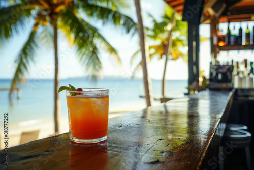 A glass of orange juice sits on a wooden bar counter next to a palm tree. The drink is garnished with a lime wedge and a cherry. The scene is set on a beach, with the ocean visible in the background