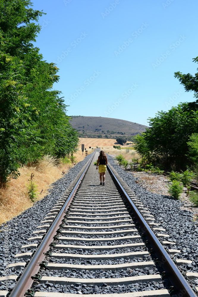 Fototapeta premium Young woman walks thoughtfully along the railroad tracks through the countryside