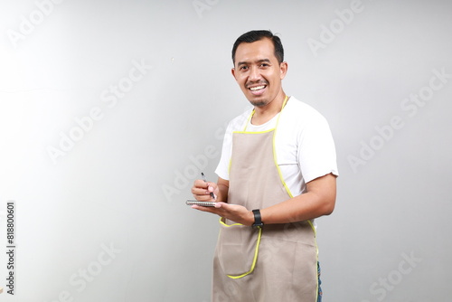 Friendly Asian barista man in brown apron taking order, writing on menu book list, looking at the camera. Isolated on gray background photo