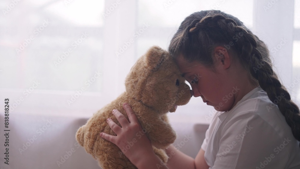 depressed girl child sitting by the window with her favorite toy teddy bear upset. problem of domestic violence in the family concept. depressed girl is sad sunlight and bored at window with bear depressed girl child sitting by the window with her favorite toy teddy bear upset. problem of domestic violence in the family concept. depressed girl is sad sunlight and bored at window with bear