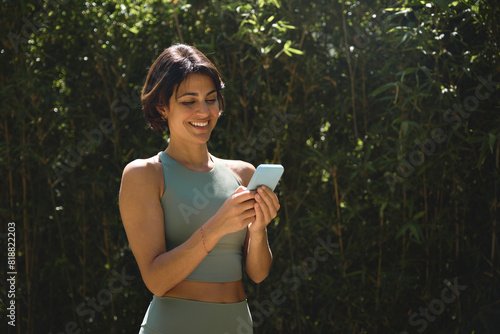 Fototapeta Naklejka Na Ścianę i Meble -  Happy Hispanic fit young woman using mobile phone checking fitness applications standing in green nature park. Online free trainings exercises, sport and health care commercial apps concept.