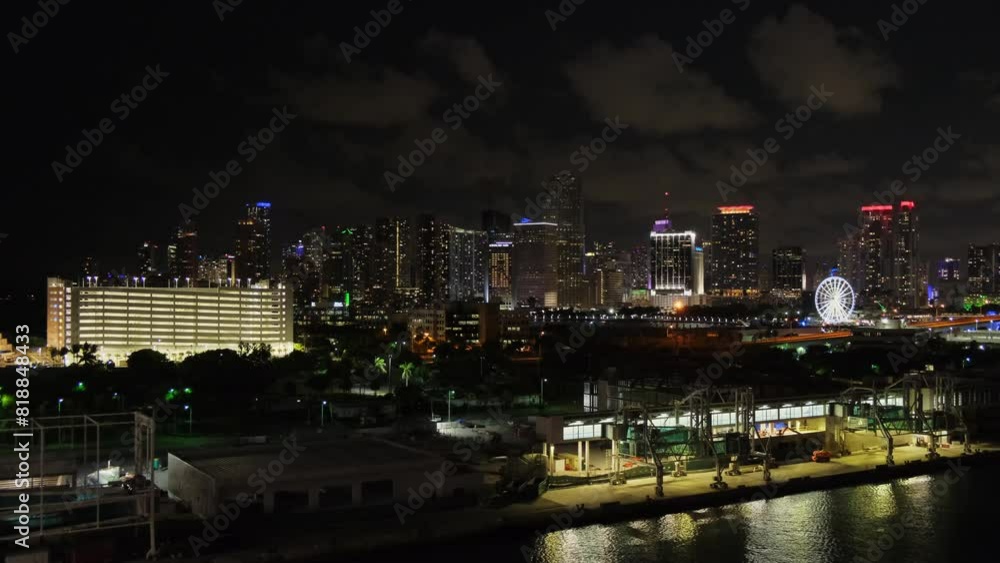 Night view from afar of Miami, Florida, from port, cruise terminal. Industrial buildings, calm water surface, silhouettes of city downtown illuminated skyscrapers in distance against dark cloudy sky. Night view from afar of Miami, Florida, from port, cruise terminal. Industrial buildings, calm water surface, silhouettes of city downtown illuminated skyscrapers in distance against dark cloudy sky.