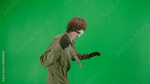 Maniac in hockey mask and jumpsuit running and waving a large sharp knife. An unrecognizable man in the terrifying guise of Jason Voorhees on a green screen chromakey. Halloween, horror. Side view.