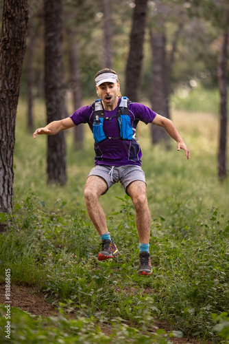 A man in a purple shirt and gray shorts is jumping in a forest. He is wearing a visor and has a backpack on