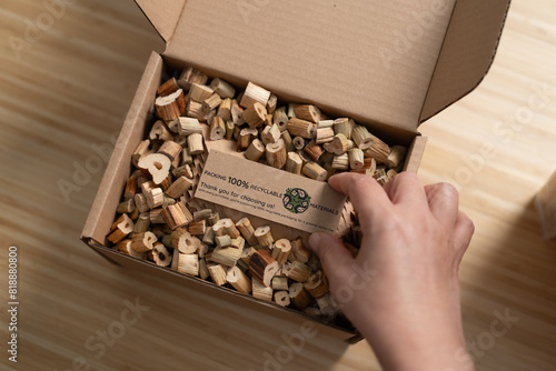 A woman places a "100% Recyclable Materials" label on a cardboard box filled with eco-friendly water hyacinth cushioning, demonstrating sustainable and environmentally friendly business practices