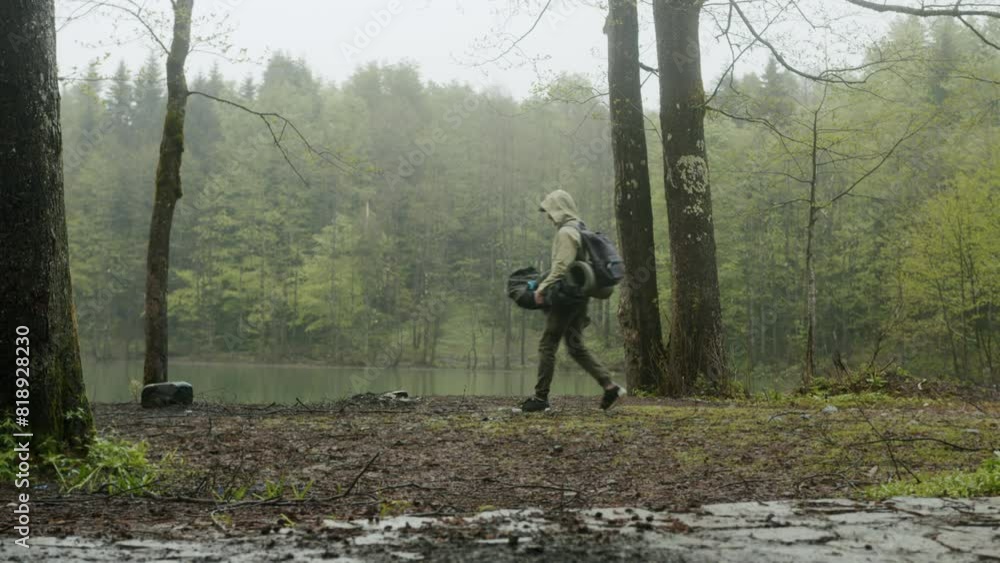 Hiker man assembles campsite tent in the rainy forest near the lake. Traveler installing tent in woods. Outdoor weekend activity. Wide shot Hiker man assembles campsite tent in the rainy forest near the lake. Traveler installing tent in woods. Outdoor weekend activity. Wide shot