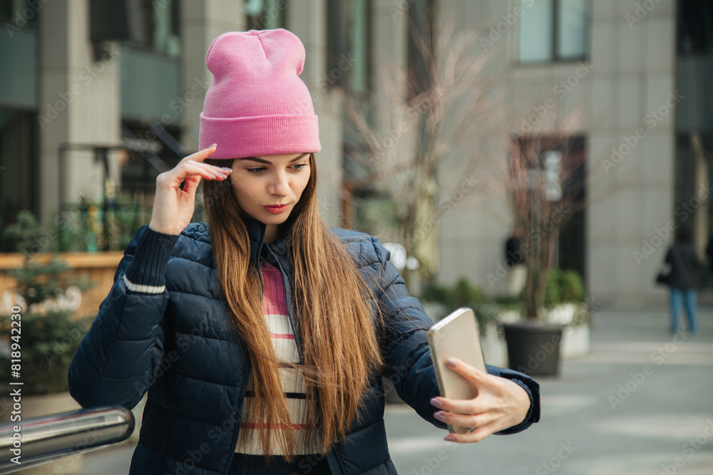 Portrait of a young woman in a pink hat and blue jacket taking a selfie on the street.