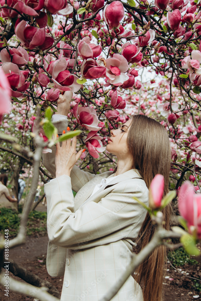 Fototapeta premium A young woman in a jacket enjoys blooming magnolia trees under a large tree.