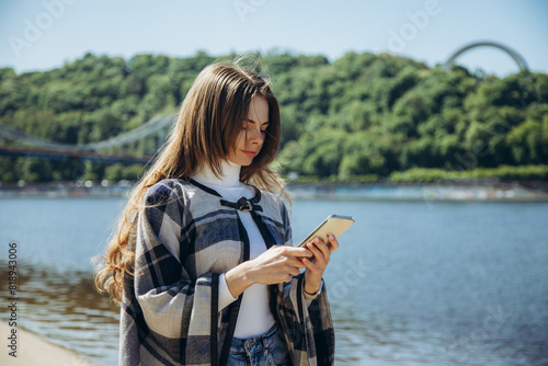 A young woman in a poncho on the bank of the river is typing a message on the phone.