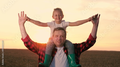 Smiling father and daughter walking at sunset agriculture wheat field closeup. Cute little girl sitting on male parent shoulders with open hands enjoy freedom happy childhood sunrise sky meadow © DREAM INSPIRATION