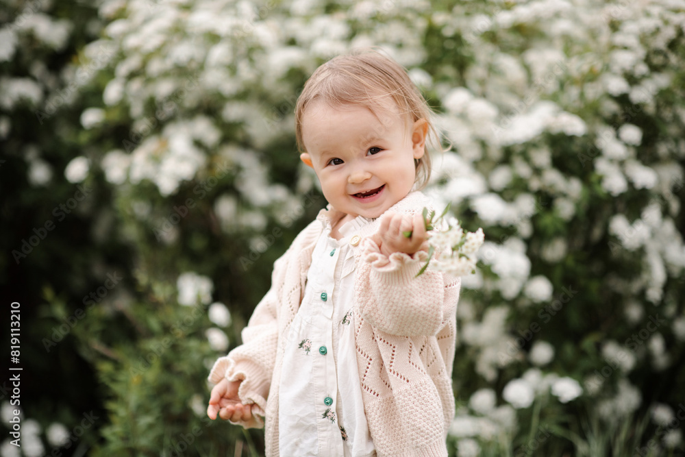 Smiling baby girl 1-2 year old wearing stylish clothes standing over blooming bushes with flowers outdoor. Looking at camera. Childhood.